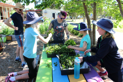 exteriror, three primary school girls planting seedlings on a multicoloured picnic bench while a male and female adult watch