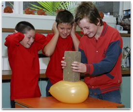 A student pushing a brick onto a balloon. Two other students are watching.