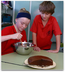Two students are mixing a cake mixture. A chocolate cake is in the foreground.