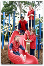 Students on play equipment, watching a girl slide down a playground slide.