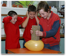 A student pushing a brick onto a balloon. Two other students are watching.