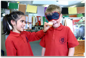 A girl is holding onion up to the nose of another blindfolded student.