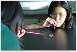 2 students are blowing through straws competing to make a large marble roll.