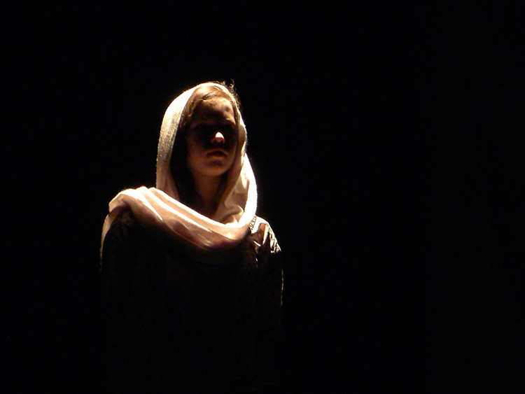 a photograph of a female actor on stage. Low key lighting is used. The background is dark, and her face is in shadow. Her shawl over her head and around her neck is lit up.