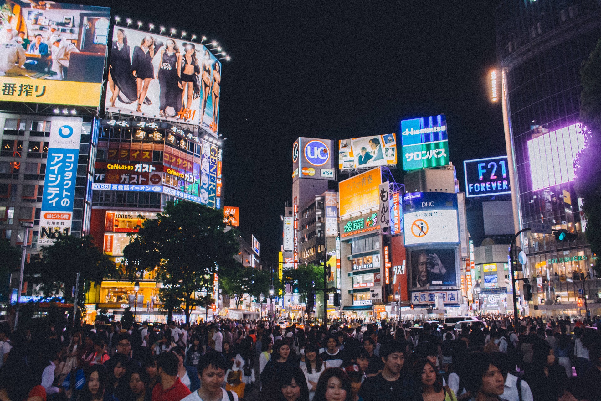 a photograph of Shibuya crossing, Tokyo, at night. The crossing is crowded with people. In the upper background are bright, digital billboards.
